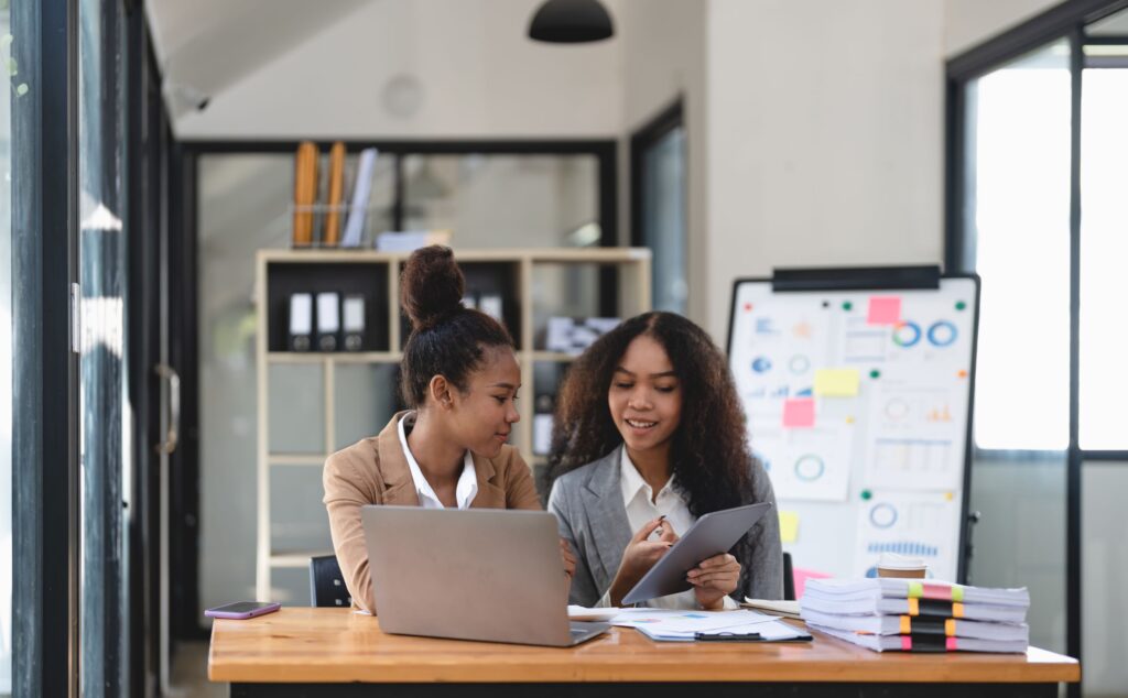 duas mulheres trabalham no computador em frente a um quadro com gráficos